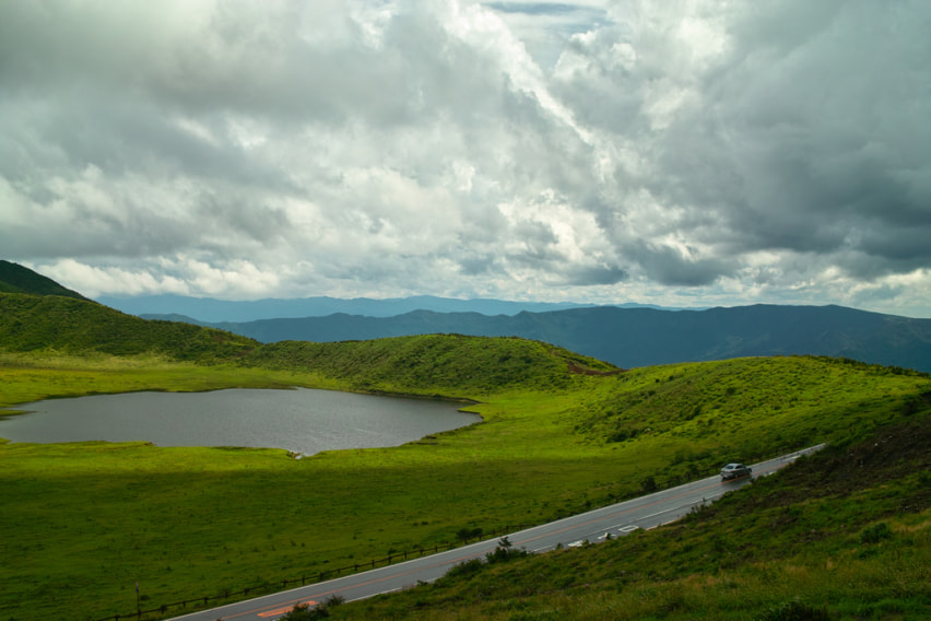 A lake and road in the middle of brilliant green hills, with clouds in the sky lit from above