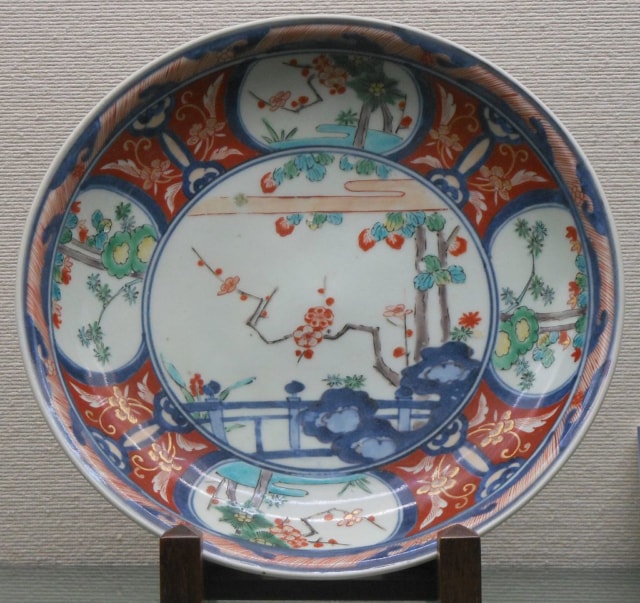 A ceramic plate on display, decorated with an intricate design in blue, red, green and pale yellow on a white background